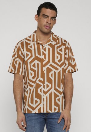 Homme portant une chemise marron à manches courtes à boutons avec un motif géométrique blanc, associée à un jean bleu, debout devant un fond clair uni.