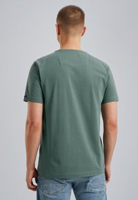 Korte mouwen T-shirt in stoffig groen katoen, met een rechte pasvorm en een rechthoekige rugpasse. Gepaarde met lichtblauwe denim jeans.