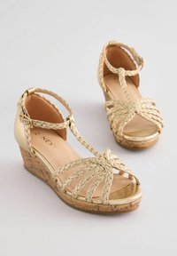 Goldene Keilsandalen mit geflochtenen Riemen und offenen Zehen. Korkähnlicher Absatz mit glattem, glänzendem Obermaterial. Verstellbarer Knöchelriemen inklusive.