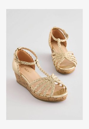 Arany wedge szandál fonott pántokkal és nyitott orral. Paraffin-szerű sarkú, sima, fényes felső anyag. Állítható bokapánttal rendelkezik.