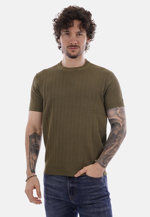 Uomo con capelli ricci che indossa una maglia a maniche corte verde oliva in maglia e jeans blu, mostra tatuaggi dettagliati su entrambi gli avambracci, in piedi contro uno sfondo bianco.