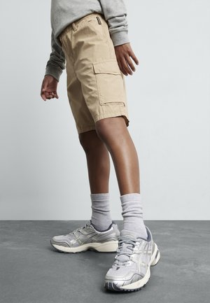 Person trägt beige Cargo-Shorts, grauen Sweatshirt, graue Socken und silberne Sportschuhe und steht auf grauem Boden.
