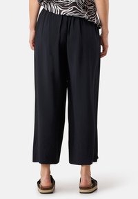 Zwarte wijde broek met een gestructureerde afwerking, voorzien van een elastische tailleband. Gepaarde met instap sandalen op een effen achtergrond.