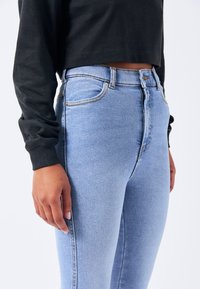 Jeans de tiro alto en azul claro con un diseño ajustado y una textura sutil. Costuras en contraste y bolsillos frontales son visibles. Top negro de manga larga.