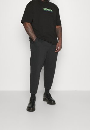 Mies, jolla on musta kuvioitu T-paita, mustat käärittyjen lahkeiden housut, mustat sukat ja mustat nahkakengät, seisoo valkoista taustaa vasten.