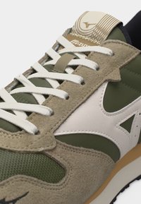 Κοντινή λήψη πράσινου και μπεζ αθλητικού παπουτσιού με λευκά κορδόνια, επενδύσεις από σουέντ και λευκό λογότυπο στο πλάι και στη γλώσσα.