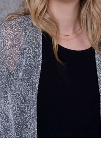 Průsvitný černobílý kardigan s paisley vzorem přes černé tričko; vrstvený design s plynulou texturou a otevřeným předním dílem. Viditelný zlatý vrstvený náhrdelník.