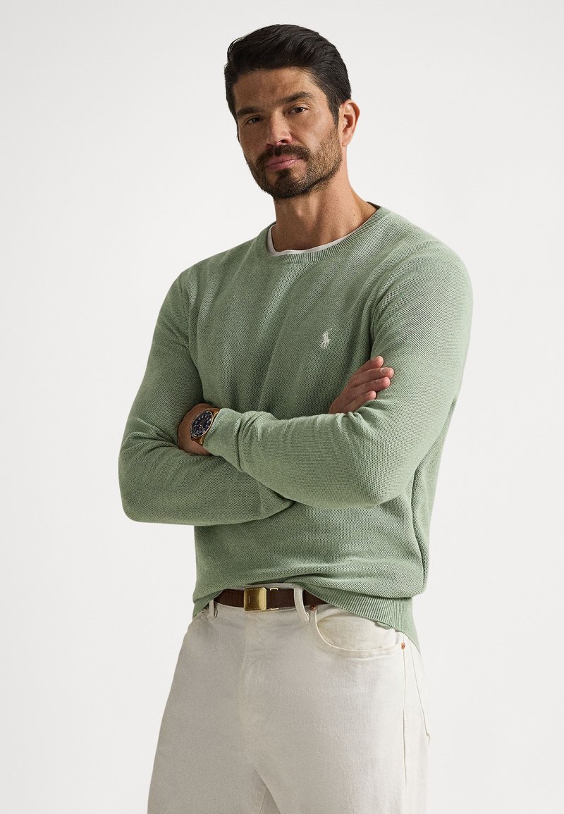 Light green knit sweater with a round neckline and ribbed cuffs, featuring a small white logo on the chest. Paired with light-colored pants.
