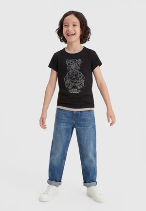 Παιδί που χαμογελά, φορώντας μαύρο μπλουζάκι με γεωμετρικό σχέδιο αρκουδάκου, μπλε τζιν με ανασηκωμένα μπατζάκια και λευκά αθλητικά, στέκεται μπροστά σε απλό φόντο.