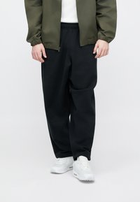 Melnas sporta bikses no mīksta auduma ar brīvu piegriezumu, elastīgu jostasvietu un sānu kabatām, kombinētas ar baltām sporta kurpēm un zaļu jaku.