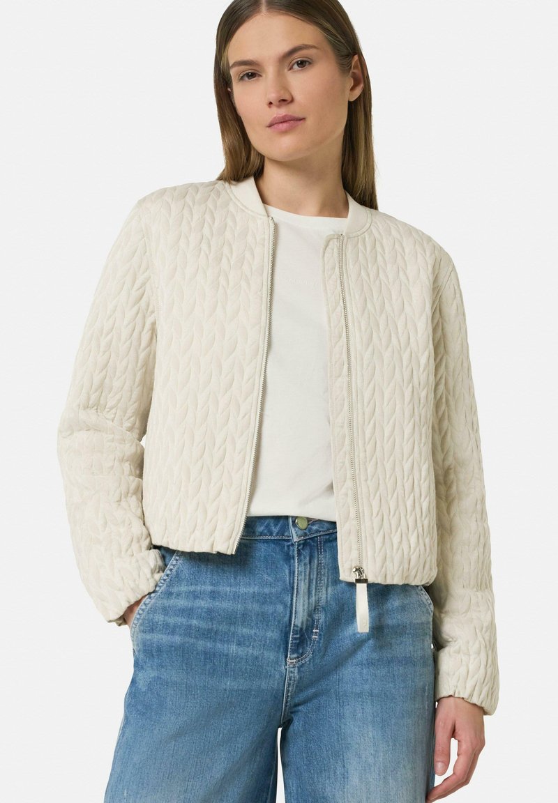 Cream quilted bomber jacket with ribbed collar and cuffs. Zipper closure at front. Worn over a white shirt and blue denim jeans.