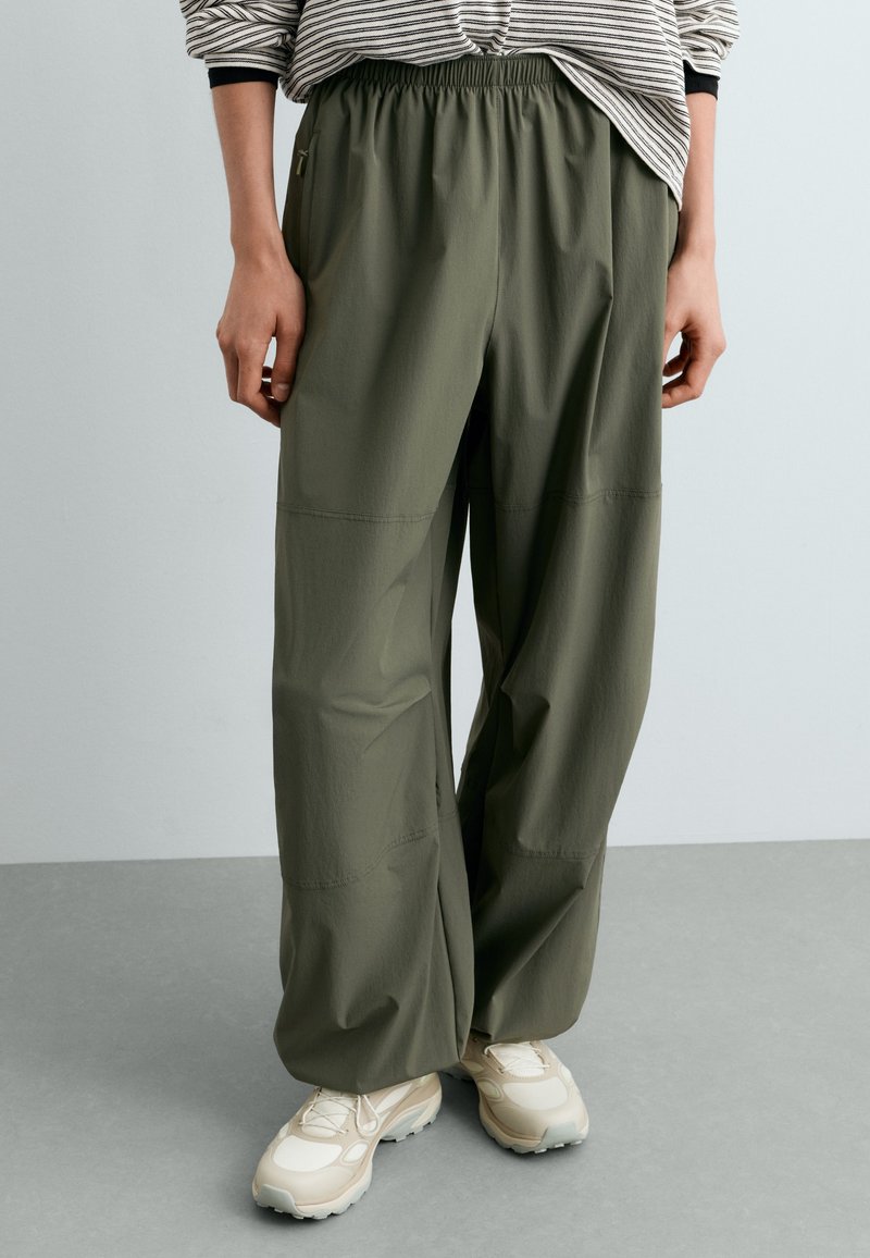Personne portant un pantalon ample vert olive avec une taille élastique et des baskets beiges, debout sur un sol gris sur fond uni.