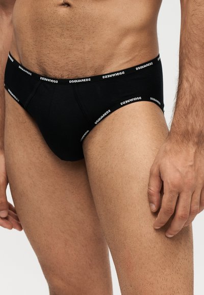 Torse et haut des jambes masculins portant un slip noir de la marque Dsquared2, main détendue sur le côté, sur un fond clair uni.