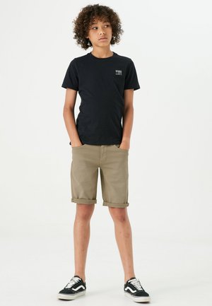 T-shirt en coton noir avec un petit logo, associé à un short en khaki retroussé. La tenue est complétée par des baskets noires et blanches.