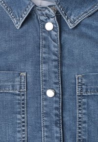 Gros plan sur le devant d'une chemise en denim bleu montrant le col, deux poches poitrine et des boutons-pression argentés avec des coutures visibles.