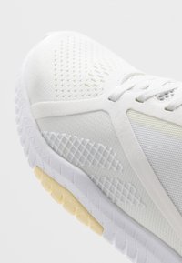 Bílá atletická obuv s horní částí z prodyšné síťoviny, texturovanými detaily a světle žlutou gumovou podrážkou. Disponuje složitými vzory a elegantním designem.