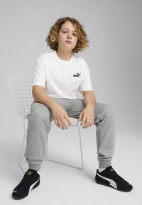 Biała koszulka z czarnym logo Puma, szare dresy i czarne sneakersy z białymi akcentami, siedzący na metalowym krześle na szarym tle.