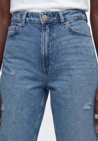 Jean en denim bleu clair taille haute avec une légère usure au niveau des cuisses, porté avec un haut blanc rentré, montrant les détails des boutons et des poches.