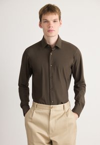 Camisa de botones marrón de manga larga con cuello puntiagudo y botones dorados, combinada con pantalones de color crema. Textura suave, corte ajustado.