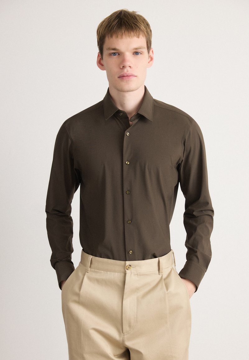 Camisa de botones marrón de manga larga con cuello puntiagudo y botones dorados, combinada con pantalones de color crema. Textura suave, corte ajustado.