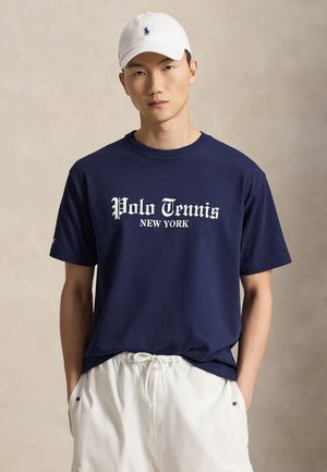 Homme portant une casquette blanche, un t-shirt bleu marine avec le texte "Polo Tennis New York" et un short blanc à cordon, debout devant un fond uni.