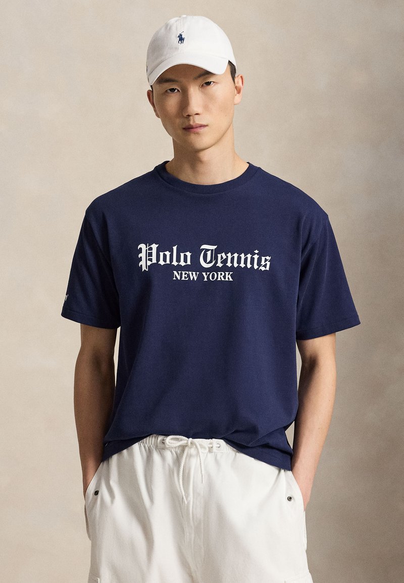 Άνδρας με λευκό καπέλο, μπλε σκούρο T-shirt με το κείμενο "Polo Tennis New York" και λευκά σορτς με κορδόνια, στέκεται μπροστά σε απλό φόντο.