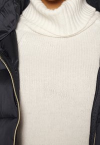 Flettet cremefarvet turtleneck sweater med ribbet tekstur, lagdelt under en sort puffjakke med en gylden lynlås.