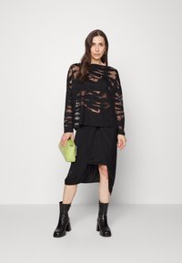 Blusa preta transparente de manga longa com recortes horizontais, combinada com uma saia preta com nó. Os acessórios incluem uma bolsa verde e botas pretas de tornozelo.