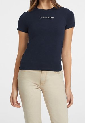 Mujer con una camiseta ajustada azul marino con el texto "GUESS JEANS" y pantalones beige, mostrada desde el cuello hasta las caderas sobre un fondo blanco.