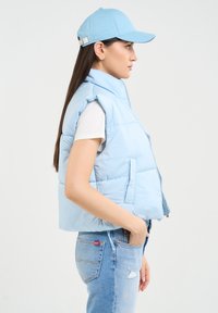 Šviesiai mėlynas pūkinis liemenė su aukštu apykakle, užtrauktuku priekyje ir šoninėmis kišenėmis, dėvimas ant baltos marškinėlių ir derinamas su šviesiai mėlyna kepure ir džinsais.