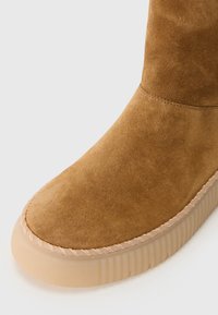Bruine suede enkellaars met een ronde neus, gestikte details en een gestructureerde, doorschijnende rubberen zool.