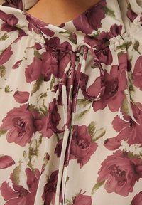 Blouse florale avec un fond crème, ornée de grands motifs de fleurs roses et bordeaux. Tissu doux avec un cordon de serrage au niveau du cou.