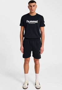 Zwart T-shirt met witte "hummel"-logo, gecombineerd met zwarte korte broek. Heeft een losse pasvorm en een elastische tailleband. Draagt witte sokken en sneakers.
