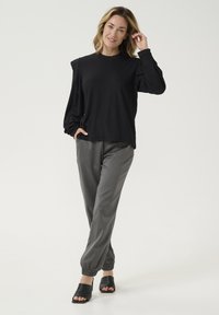Bluză neagră cu mânecă lungă, cu un design texturat, asortată cu pantaloni gri tapered, cu manșete elastice și saboți negri cu vârf deschis.