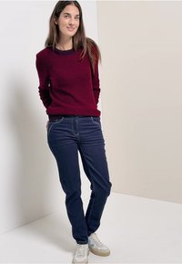 Pull rouge texturé avec un col rond, associé à un jean bleu foncé et des baskets blanches. Le modèle se tient devant un fond neutre.
