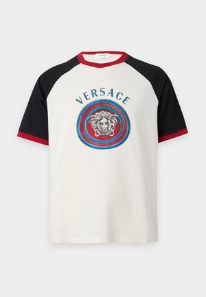Homme portant un t-shirt manches courtes avec le logo Versace, bordé de rouge et noir, un pantalon large rayé, des baskets, et portant un sac à épaule noir.