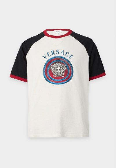 Μπλούζα Versace με κοντά μανίκια, λευκό μπροστινό μέρος, μαύρα μανίκια, κόκκινο περίγραμμα και λογότυπο κεφαλής Μέδουσας μέσα σε κόκκινο και μπλε κύκλο στο στήθος.