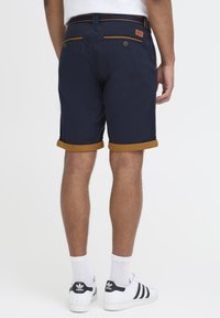 Mørkeblå shorts med oppbrettede beige mansjetter, med en glatt tekstur, to baklommer med knapper og et merkestykke i lær på livlinjen.