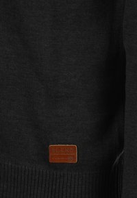 Svart stickad tröja med ribbat nederkant och ärmslut. Har en liten brun läderetikett med texten "BLEND GLOBAL JEANMAKER" på den nedre framsidan.