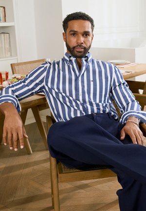 Bărbat cu păr scurt și creț și barbă, purtând o cămașă în dungi albastre și albe și pantaloni închiși la culoare, stă relaxat pe un scaun de lemn la masa de dining.
