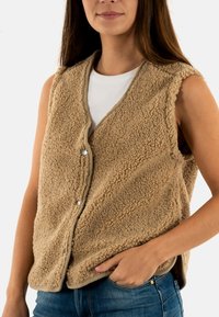 Gilet senza maniche di colore beige, realizzato in un materiale morbido e texture. Presenta un design a V e una chiusura a scatto davanti. Indossato sopra una maglietta bianca.