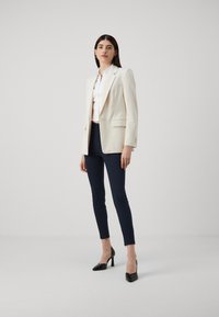 Cremeblazer med klipp-lapper, tilpasset design og frontlommer, kombinert med marineblå smale bukser og svarte høye heler. Glatt stofftekstur.