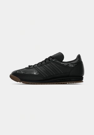 Zapatilla de cuero negra de Adidas con detalles bordados en blanco, cordones negros, suela de goma marrón y las tres franjas características en el lateral.