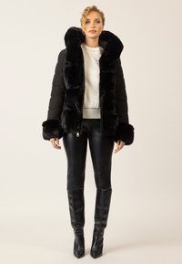 Frau in schwarzer Daunenjacke mit Pelzbesatz, weißem Pullover, schwarzen Lederhosen und knielangen schwarzen Stiefeln, die vor einem schlichten Hintergrund steht.