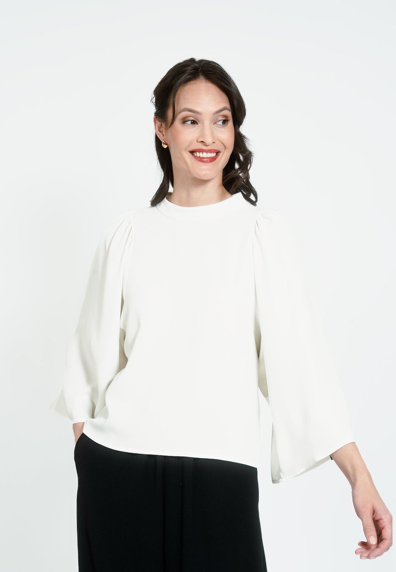 Biela blúzka s dlhou rukávou, vysokým golierom a ryšavými ramenami. Nosí sa s čiernymi nohavicami, ktoré majú hladkú textúru a voľný strih.