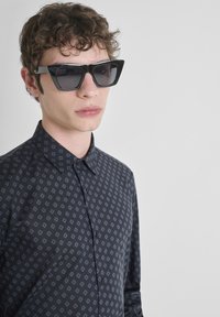 Occhiali da sole neri con montatura spessa, indossati con una camicia scura a fantasia caratterizzata da piccoli disegni a forma di diamante grigi.