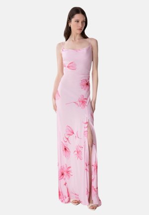 Donna che indossa un vestito rosa senza maniche, lungo fino al pavimento, con spalline sottili e stampe floreali rosse, caratterizzato da uno spacco alto sul lato sinistro.