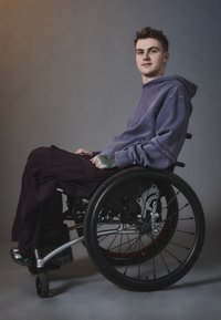 Scaun cu rotile cu un cadru argintiu elegant, roți din cauciuc negre și accente roșii; o persoană îmbrăcată cu hanorac violet și pantaloni închiși la culoare, așezată confortabil.