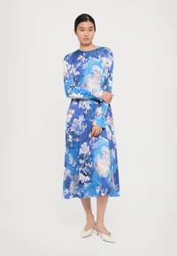 Femeie îmbrăcată într-o rochie midi cu mânecă lungă, florală, de culoare albastră, purtând pantofi cu toc mic, albi, pe un fundal simplu.