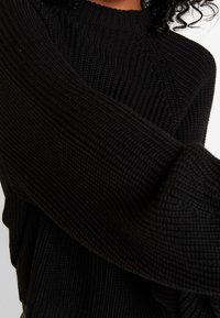 Pull noir en maille avec texture côtelée et col haut, doté d'une coupe ample et d'épaules tombantes, mettant en valeur les détails de fil épais.